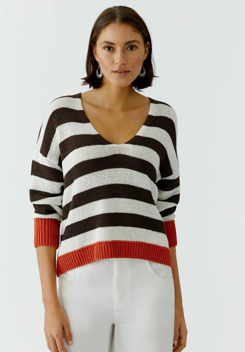 Femme portant un pull en tricot rayé marron et blanc avec des poignets et un ourlet orange, associée à un pantalon blanc, debout devant un fond blanc.