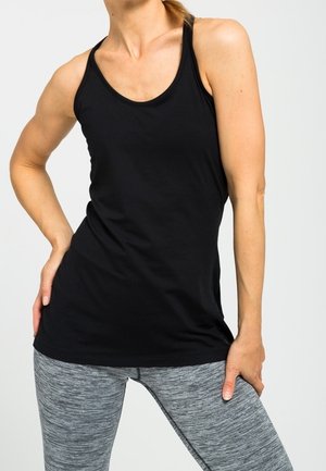 Kobieta ubrana w czarny top na ramiączkach typu racerback i szare legginsy w wzory, z jedną ręką na biodrze, a drugą opartą na udzie.