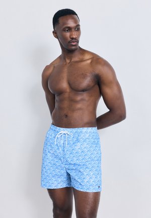 Muskularny mężczyzna o ciemnej karnacji, ubrany w niebieskie kąpielówki z wzorem, stoi na jasnoszarym, jednolitym tle z rękami za plecami.