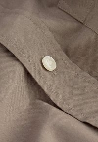 Un tejido marrón con una textura fina presenta un gran botón blanco y detalles cosidos. El enfoque está en la superficie suave del botón y el hilo.