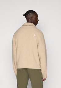 Maglione beige a costine con colletto sciallato, caratterizzato da un dettaglio logo bianco sulla schiena, abbinato a pantaloni verde oliva.