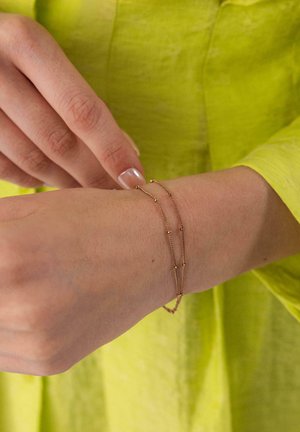 Hand wearing yellow sleeve adjusting delicate double gold chain bracelet with small beads on wrist.