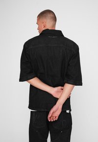 Zwarte denim overhemd met een gestructureerde kraag en korte mouwen, voorzien van een subtiel logo op de achterkant en een textuur stof. Ontspannen pasvorm ontwerp.