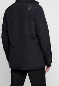 Takapään näkymä henkilöstä, joka on pukeutunut mustaan topattuun takkiin, jossa on "Cars Jeans 1982" brodeerattu olkapään lähelle ja mustiin housuihin.