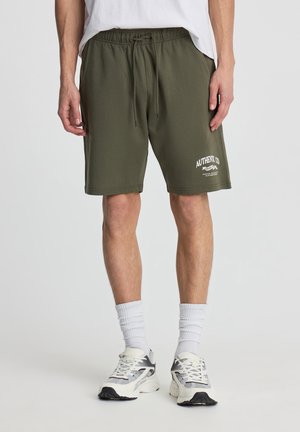 Persona che indossa pantaloncini verde oliva con coulisse e logo bianco sulla gamba sinistra, calzini bianchi e scarpe sportive bianche e grigie.