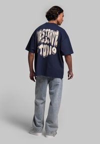Navyblauwe oversized t-shirt met witte grafische tekst op de achterkant; gedragen met lichtblauwe denim jeans en witte sneakers.