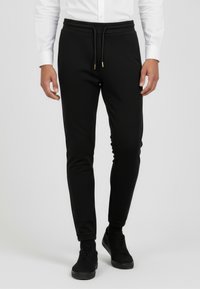 Schwarze Slim-Fit-Jogginghosen aus weichem Stoff, mit einem Kordelzugbund mit goldenen Akzenten und zulaufenden Beinen. Getragen mit schwarzen Turnschuhen.