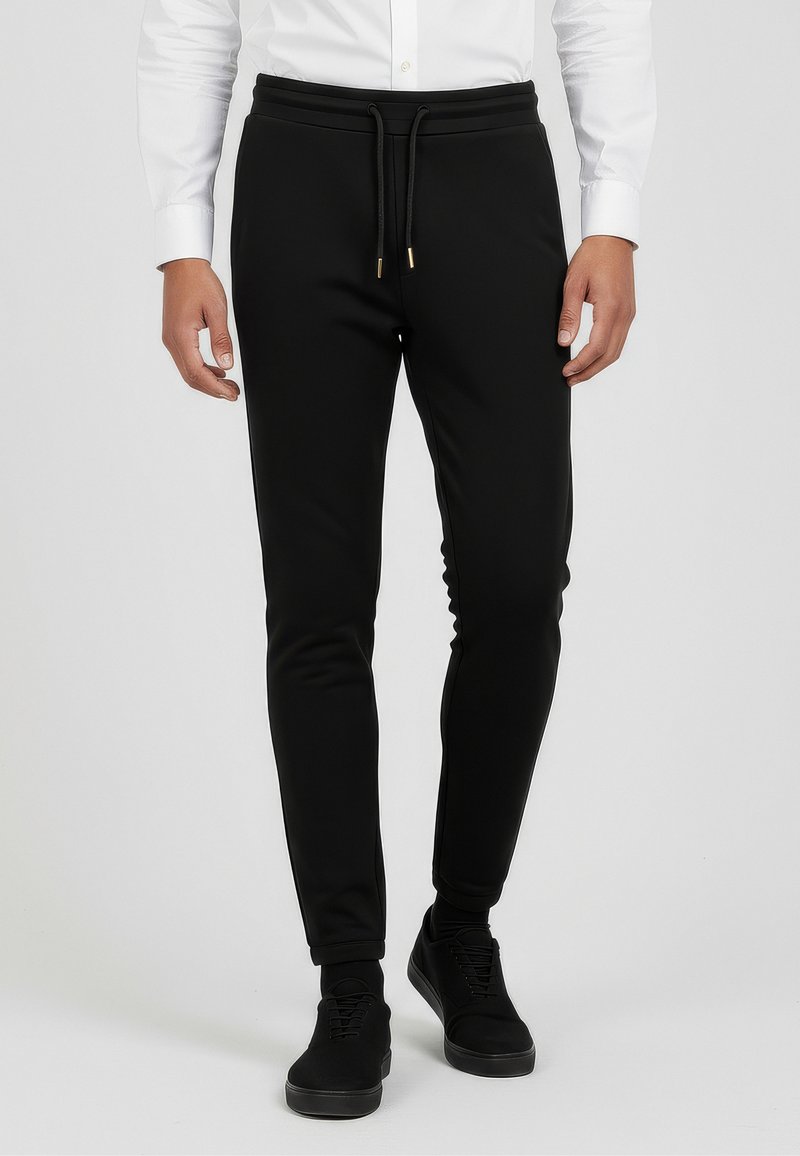Schwarze Slim-Fit-Jogginghosen aus weichem Stoff, mit einem Kordelzugbund mit goldenen Akzenten und zulaufenden Beinen. Getragen mit schwarzen Turnschuhen.