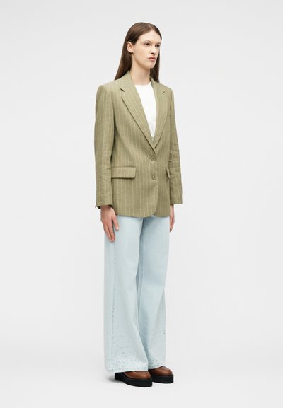 Femme aux longs cheveux bruns portant un blazer rayé olive clair, un haut blanc, un jean bleu clair à jambes larges et des chaussures à plateforme marron, debout.