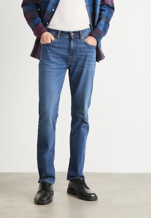 Jeans blu scuro con vestibilità dritta, leggera scoloritura, cinque tasche, indossati con una maglietta bianca e scarpe nere stringate. Camicia a quadri appoggiata.