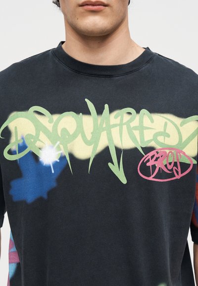 Κοντινό πλάνο ενός ατόμου που φοράει μαύρο μπλουζάκι με κείμενο και σχέδια σε στυλ γκράφιτι σε πράσινο, κίτρινο, μπλε και κόκκινο στο στήθος.