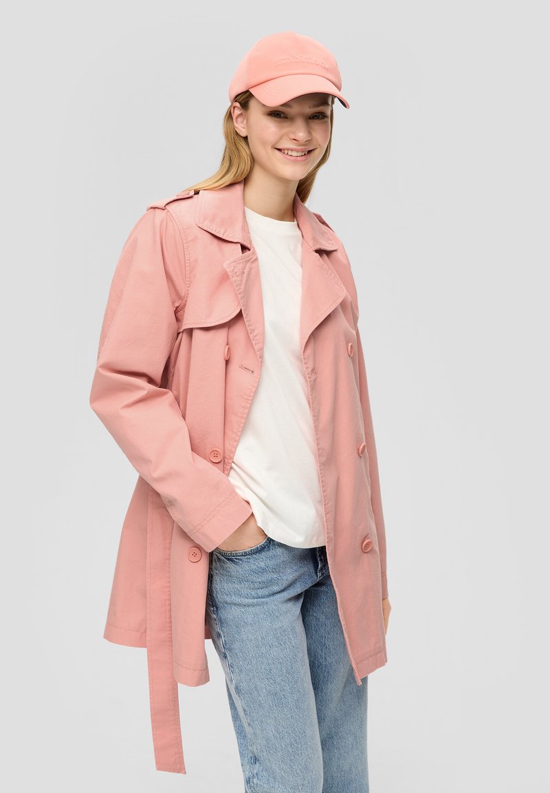 Lichtroze trenchcoat met een dubbele knopenontwerp, brede kraag en strikriem. Gekocht over een wit shirt en gecombineerd met blauwe jeans.