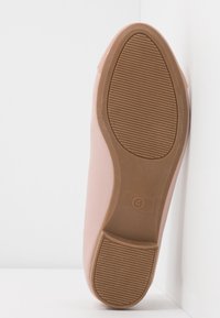 Semelle d'une chaussure plate rose clair avec une semelle en caoutchouc marron et la taille 4 marquée sur la zone du talon, positionnée verticalement contre un coin blanc.