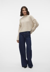 Beige strikket genser med ribbestruktur og sideskjæringer, kombinert med mørkeblå vide jeans og svart spissede sko.