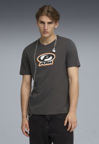 Grå kortærmet T-shirt med rund halsudskæring og et orange og sort Puma-logo i midten. Båret med sorte jeans og hovedtelefoner.