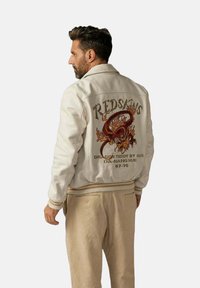 Veste bombardier blanche avec des poignets côtelés. L'arrière présente un design coloré de dragon et le texte "REDSKINS DRAGON TEDDY BY RDS IDN-MANG HUE 67-70."