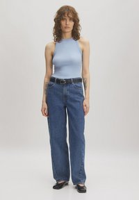 Hellblaues ärmelloses Oberteil mit hohem Kragen, kombiniert mit hochgeschnittenen blauen Jeans und einem schwarzen Gürtel. Dunkle Schuhe runden das Outfit ab.