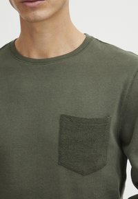 Olivengrønn langermet skjorte med en teksturert lomme. Har en rund hals og mykt, glatt stoff. Enkel design uten dekorasjoner.