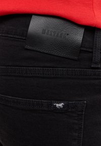 Czarne dżinsy denimowe z skórzaną metką MUSTANG przy pasku oraz małym białym logo konia na kieszeni, charakteryzujące się gładką fakturą.