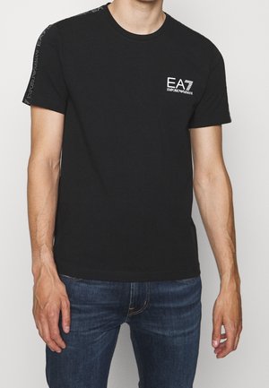 Homme portant un t-shirt noir Emporio Armani EA7 avec logo sur la poitrine gauche et texte de marque le long de l'épaule droite, associé à un jean bleu.