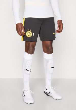 Mannlig utøver iført svarte Borussia Dortmund-shorts med logo, hvit langermet skjorte, hvite leggskinn og hvite fotballsko på grått gulv.