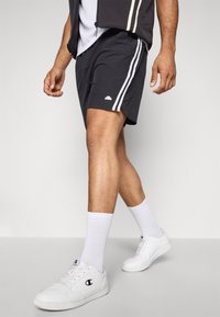 Pantalones cortos deportivos negros con rayas blancas en los laterales, combinados con zapatillas blancas y calcetas cortas. Tejido suave, ajuste casual, detalle de logo discreto.