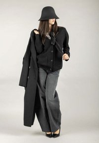 Cappotto di lana nero, cardigan nero oversize con tasche, pantaloni larghi grigi e un cappello cloche nero. Tacchi a punta neri completano il look.