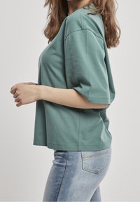 Femme portant un t-shirt vert ample à manches courtes et un jean en denim bleu clair, debout de profil avec les mains légèrement jointes devant elle.