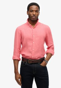 Lichtroze linnen overhemd met een staande kraag, knoopsluiting aan de voorkant en opgerolde mouwen. Draag het met donkerblauwe jeans en een bruine riem.