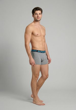 Ung mand stående barfodet iført grå Superdry boxershorts med blå taljebånd, der ser fremad mod en ensfarvet grå baggrund.