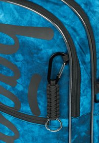 Zaino blu tie-dye con hardware nero, dotato di maniglia in paracord, grande logo, texture liscia e una zip prominente.