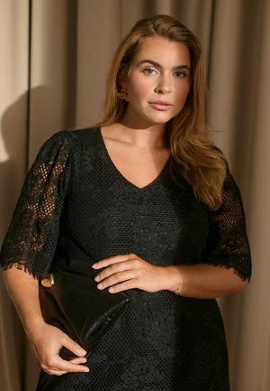 Frau mit langen hellbraunen Haaren, die ein schwarzes Spitzenkleid trägt und eine schwarze Leder-Clutch vor einem beigen Vorhanghintergrund hält.