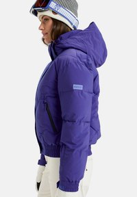 Violetti puffatakki, jossa on huppu, mustilla vetoketjullisilla taskuilla, joustavilla hihansuilla ja logopatchilla hihassa. Suuri, lämpöeristetty rakenne.