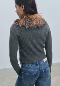 Sivkasta rebraste jopico s umetnim krznom v odtenkih rjave in sive, v kombinaciji z modrimi kavbojskimi hlačami z zadnjimi žepi.