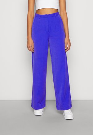 Femme portant un pantalon large bleu vif, un top court blanc et des baskets blanches, debout devant un fond clair uni.