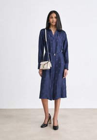 Abito satinato blu navy con colletto, maniche lunghe, tasche laterali e vita cinturata; abbinato a una piccola borsa crema e tacchi neri.