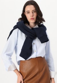 Un sweat-shirt marine noué autour du cou, porté sur une chemise à boutons bleu clair avec des boutons dorés et une jupe en cuir marron avec des détails sur les coutures latérales.