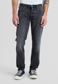 Mand iført falmede sorte jeans og sorte lave sneakers med hvide snørebånd, stående mod en ensfarvet grå baggrund.