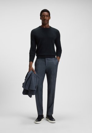 Pullover de malha escura, calças de padrão xadrez cinza claro sob medida e sapatilhas pretas. Segurando um blazer combinado, em pé contra um fundo liso.