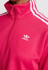 Różowa kurtka zapinana na zamek, wykonana z gładkiego materiału, z białymi paskami na ramionach i małym białym logo Adidasa na piersi.