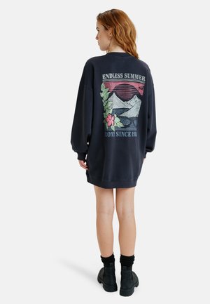 Übergroßes navy Sweatshirtkleid mit einem grafischen Design, das Berge, Blumen und Text auf der Rückseite zeigt. Aus Baumwollmaterial mit langen Ärmeln.