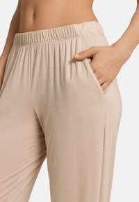 Beige, kevytrakenteiset kangashousut, joissa on joustava vyötärö ja sivutaskut. Sujuva rakenne, jossa on hienovarainen kiilto, suunniteltu mukavuutta ja helppoutta silmällä pitäen.