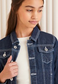 Temno denim jakna z belim šivom, dvema žepoma na prsih in srebrnimi gumbi. Nosi se čez rebrast svetel zgornji del.