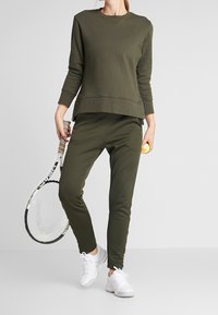 Sudadera verde oliva de manga larga y cuello redondo, combinada con joggers ajustados a juego y zapatillas deportivas blancas.
