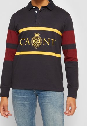 Mann trägt navyfarbenes langärmeliges Polo mit gelben Streifen und GANT-Logo, kombiniert mit hellblauen Jeans, steht vor einfachem Hintergrund.
