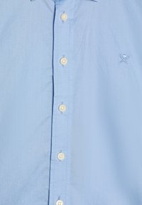 Ljusblå skjorta med knappar i vitt och en liten broderad logga med korslagda golfklubbor och boll på bröstet.