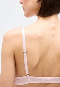 Γυναίκα με κοντά μαλλιά που φοράει ροζ ανοιχτό σουτιέν, από πίσω, με εμφανή τον ιμάντα στον ώμο και το κλείσιμο με αγκράφα.