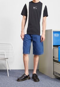 Schwarz-weißes Nike-Logo-T-Shirt, blaue Denim-Shorts und schwarze Slipper mit gelben Akzenten. Einfache Kulisse mit einem Aktenschrank.
