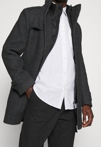 Manteau gris foncé texturé en tissu tissé, doté d'un col montant, de fermetures à pression et d'une doublure intérieure, porté par-dessus une chemise blanche.