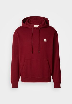 Vyno raudonos spalvos gobtuvinė sweatshirt, pagaminta iš minkštos medvilnės, su priekiu kišene, virvelėmis ir mažuoju logotipo pleistru, taip pat su gumuotais rankogaliais ir apatine dalimi.